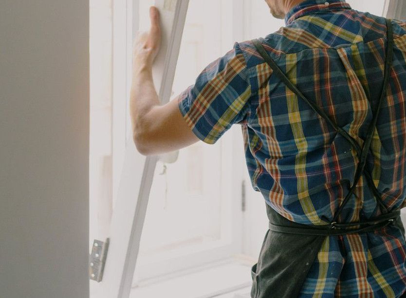 Back view of anonymous male fitter in checkered shirt and apron mounting window with hinges near white walls at home